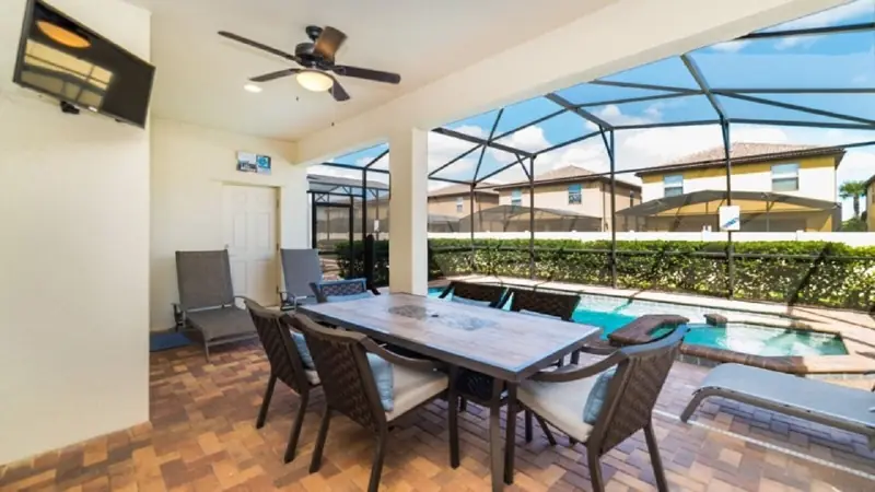 Villa lanai, pool, spa, TV and patio – A spacious patio with a wooden table and chairs, a ceiling fan, and a view of a swimming pool and neighboring houses.