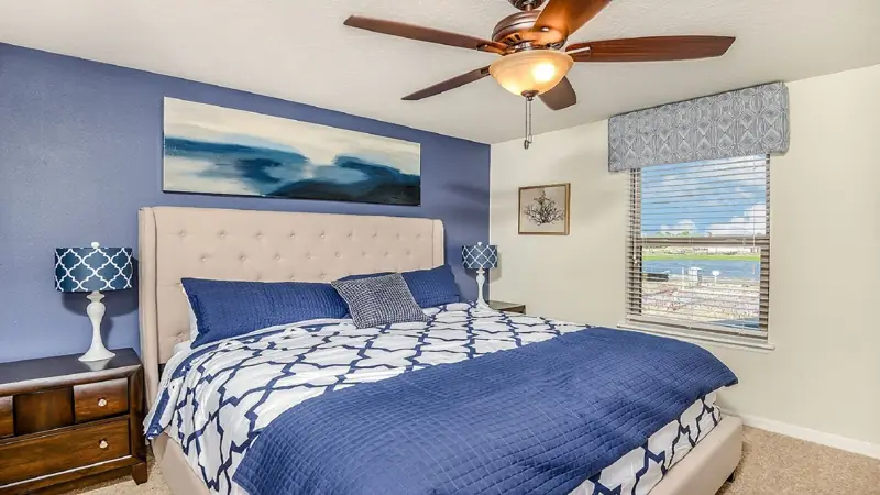 2nd floor king – A bedroom with a bed, two bedside tables, a ceiling fan, a window with blinds, and a view of a body of water.